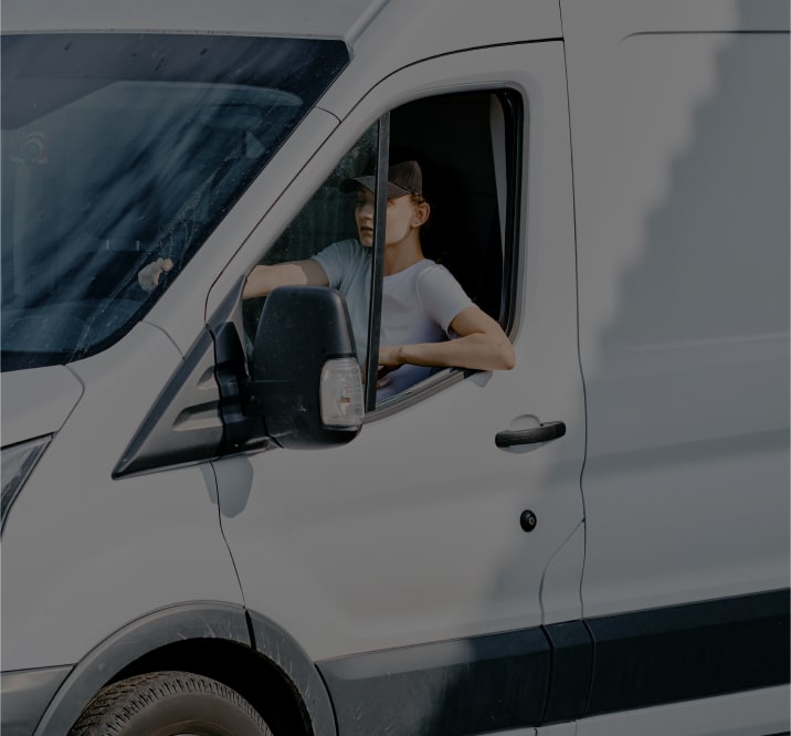 Young man wearing a white teeshirt and a dark cap driving a white van with the window down.
