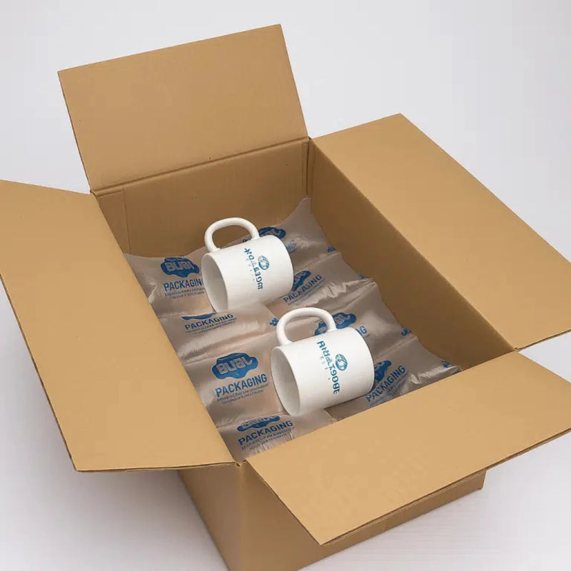 Two mugs in a cardboard box lying on a layer of protective Bubl Void 20x20 air cushioning on a white background