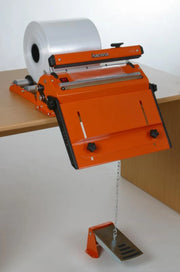 A Hacona S420 Heat Sealer mounted on a wooden table, with a clear plastic roll loaded into the sealing mechanism and a metal foot pedal connected below by a chain.