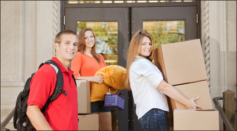 Three students with cardboard boxes and backpacks in front of a building