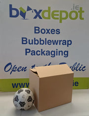 A plain brown cardboard box standing upright on a white surface, with a black-and-white soccer ball in front of it and a large sign advertising Box Depot