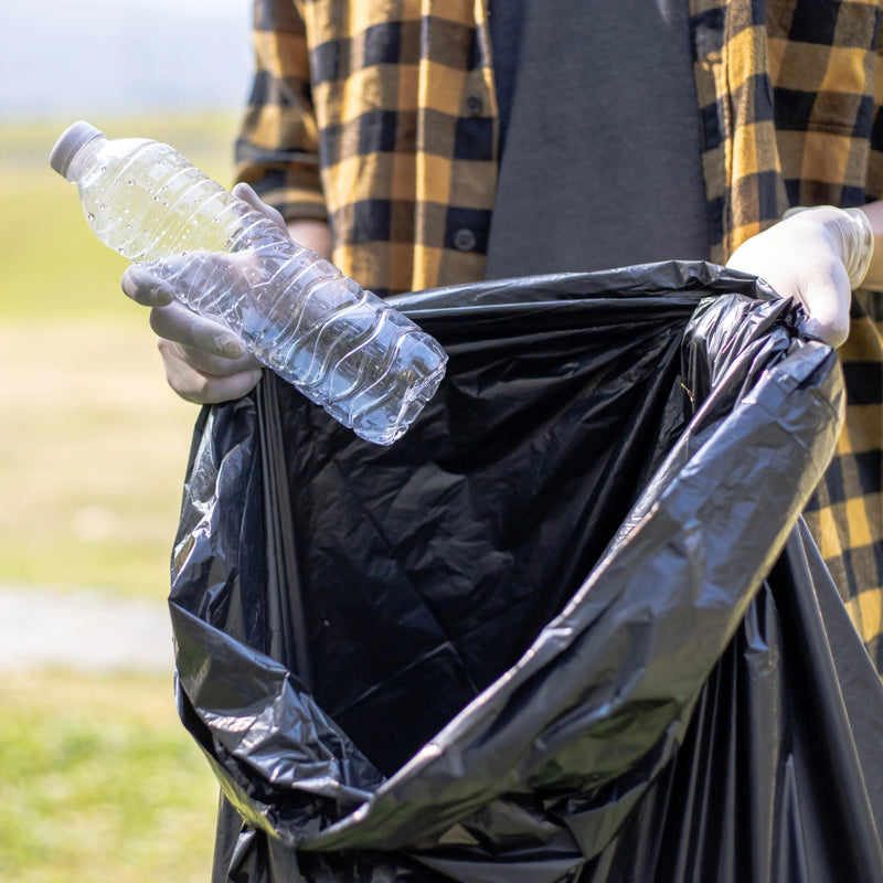Man holding a open black bin bag, putting an empty plastic bottle into it in an outdoor environment