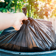 Hand holding a black refuse bag over a blue bin with a blurred green outdoor background
