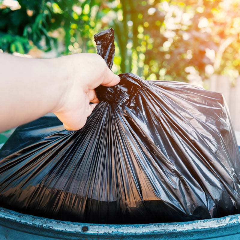 Hand holding a black refuse bag over a blue bin with a blurred green outdoor background