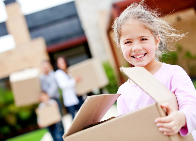 Child holding a cardboard box with her parent and moving boxes in front of their new house in the background