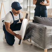 A man wrapping a fabric sofa with clear pallet wrap in an empty living room