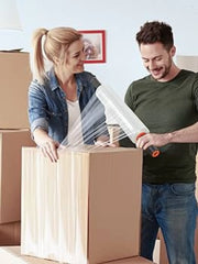 two people wrapping a cardboard box with clear pallet wrap surrounded by other cardboard boxes in a living room