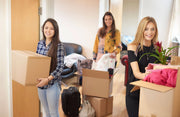 Three young women moving house, each holding a cardboard box surrounded by moving boxes in a living room setting.