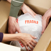 Person handling a package wrapped in bubble wrap and sealed with fragile tape inside a cardboard box.