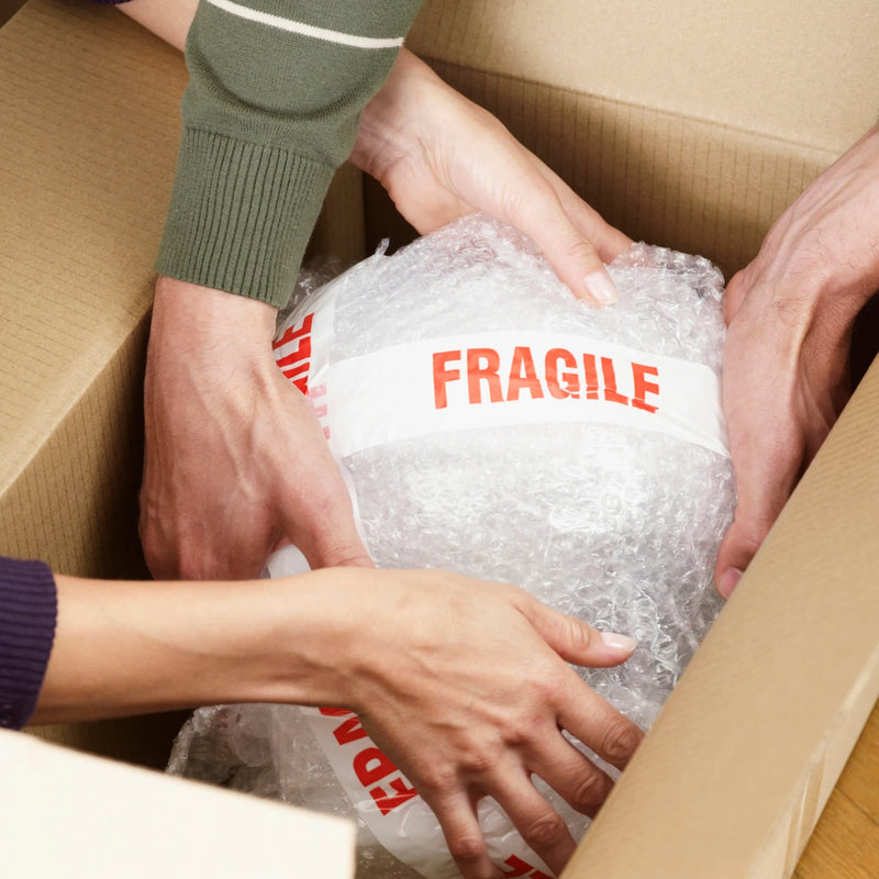 Person handling a package wrapped in bubble wrap and sealed with fragile tape inside a cardboard box.