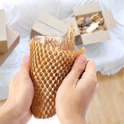 hands wrapping a glass in protective honeycomb paper with other items in boxes in the background that have also been wrapped in honeycomb paper