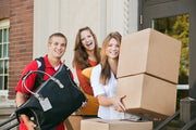 three college students standing in from of a college or university entrance holding moving boxes and bags.