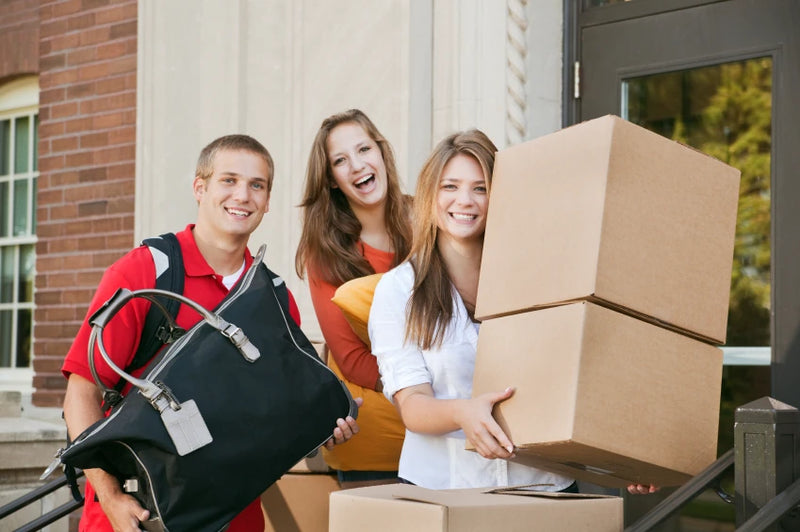 three college students standing in from of a college or university entrance holding moving boxes and bags.