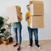 Two people holding cardboard boxes in a room with houseplants behind them.