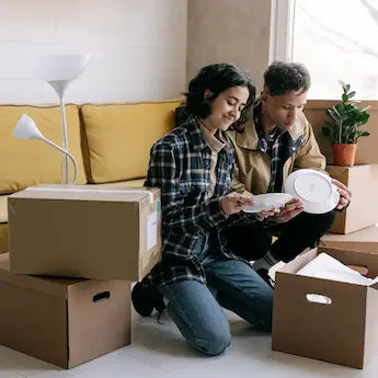 A couple sitting on the livingroom floor surrounded by cardboard boxes, and packing up plates and other belongings