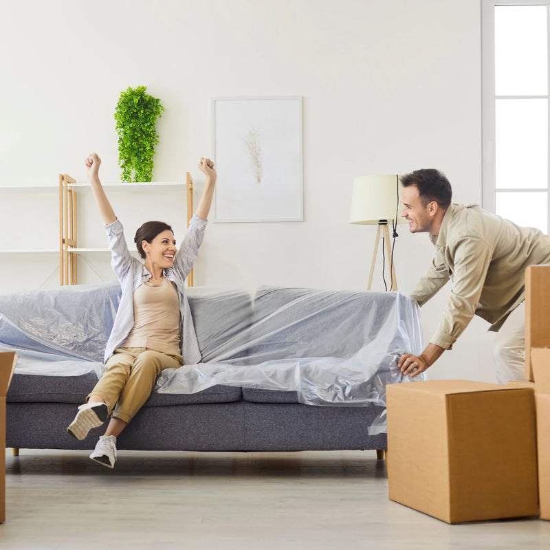 Woman sitting on a sofa with covered in a furniture cover bag, man standing nearby with moving boxes.