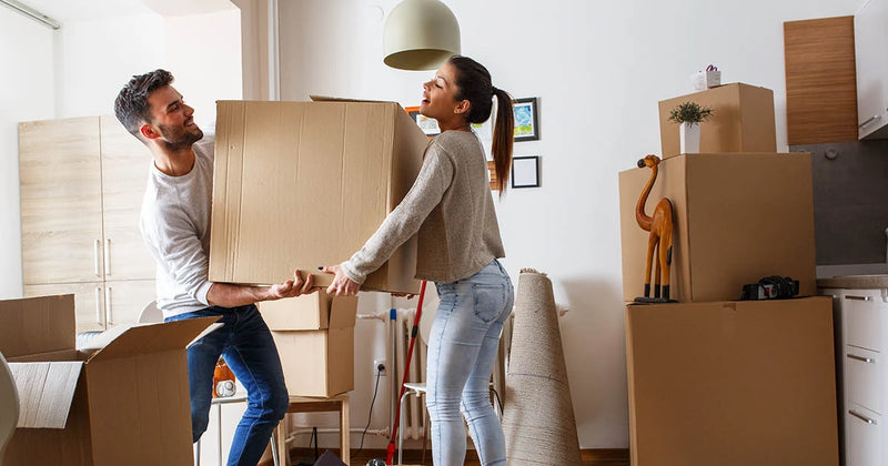 Man and woman carrying a large cardboard box with unpacked moving boxes in a room with furniture and some home decor.