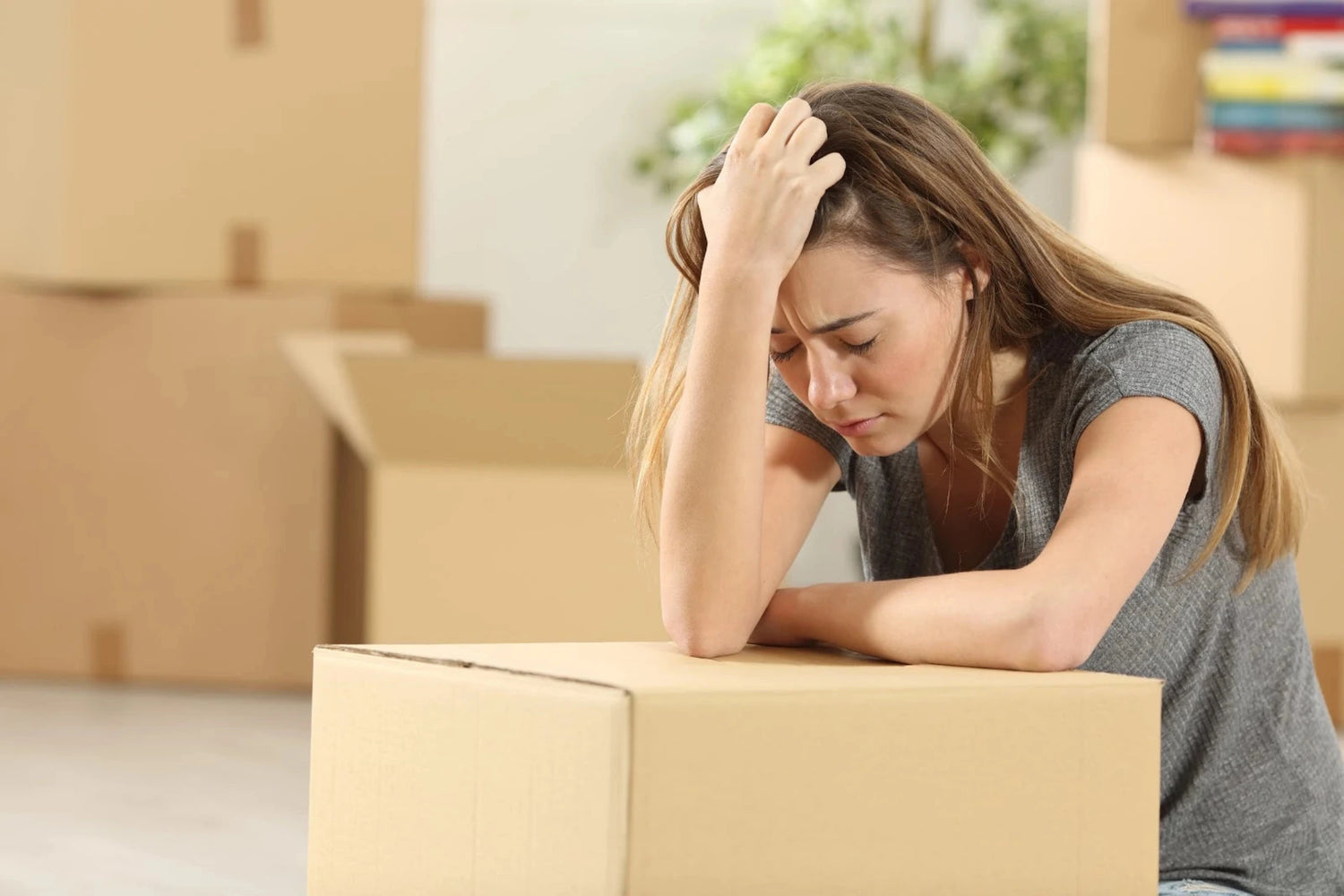 Woman sitting on a cardboard box in a room with moving boxes and books. Her face suggests frustration and procrastination at moving house.
