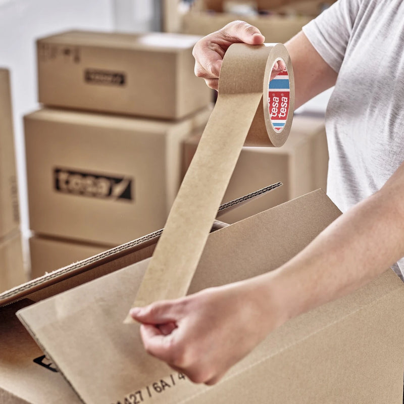 a man taping a cardboard box closed using brown paper tape in a warehouse environment