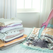 Person using a vacuum cleaner to remove air from a vacuum storage bag  next to folded clothes on a bed.