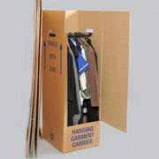 A wardrobe box with clothes on hangars on the rail of the wardrobe box with some unopened cardboard wardrobe boxes leaning on it on a white background