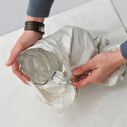 Person wrapping a glass jug with white packing paper on a light grey background