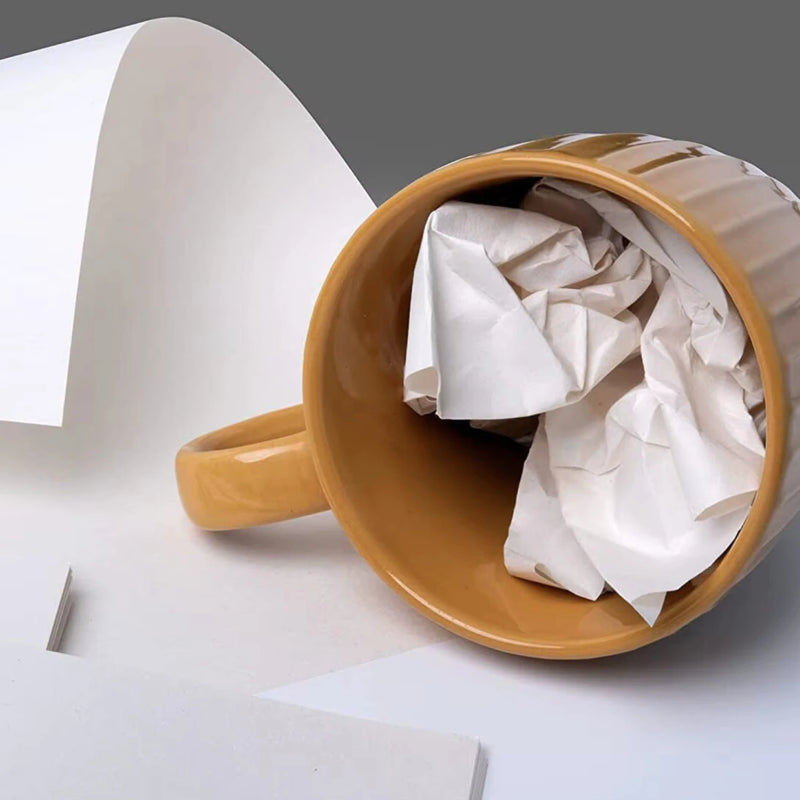 Brown mug with crumpled white packing paper inside it, lying on its side on white packaging paper on a light grey background