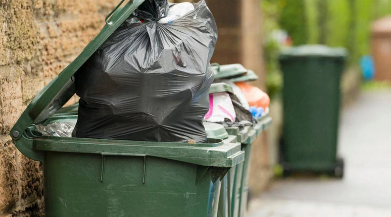 Green recycling bin with overflowing bin bags and additional items on a path at the side of a road